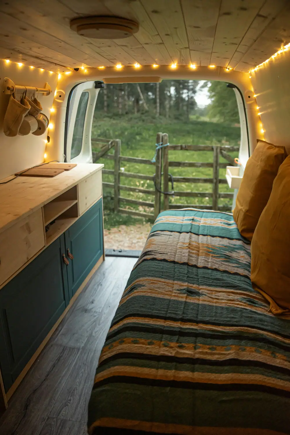 Innenraum eines kleinen Campervans mit gemachtem Bett, Holzschränken und offener Schiebetür zum Feld